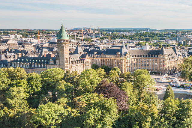Aerial view of Luxembourg City featuring the historic State Savings Bank building with its iconic tower, surrounded by lush green trees and urban landscape.